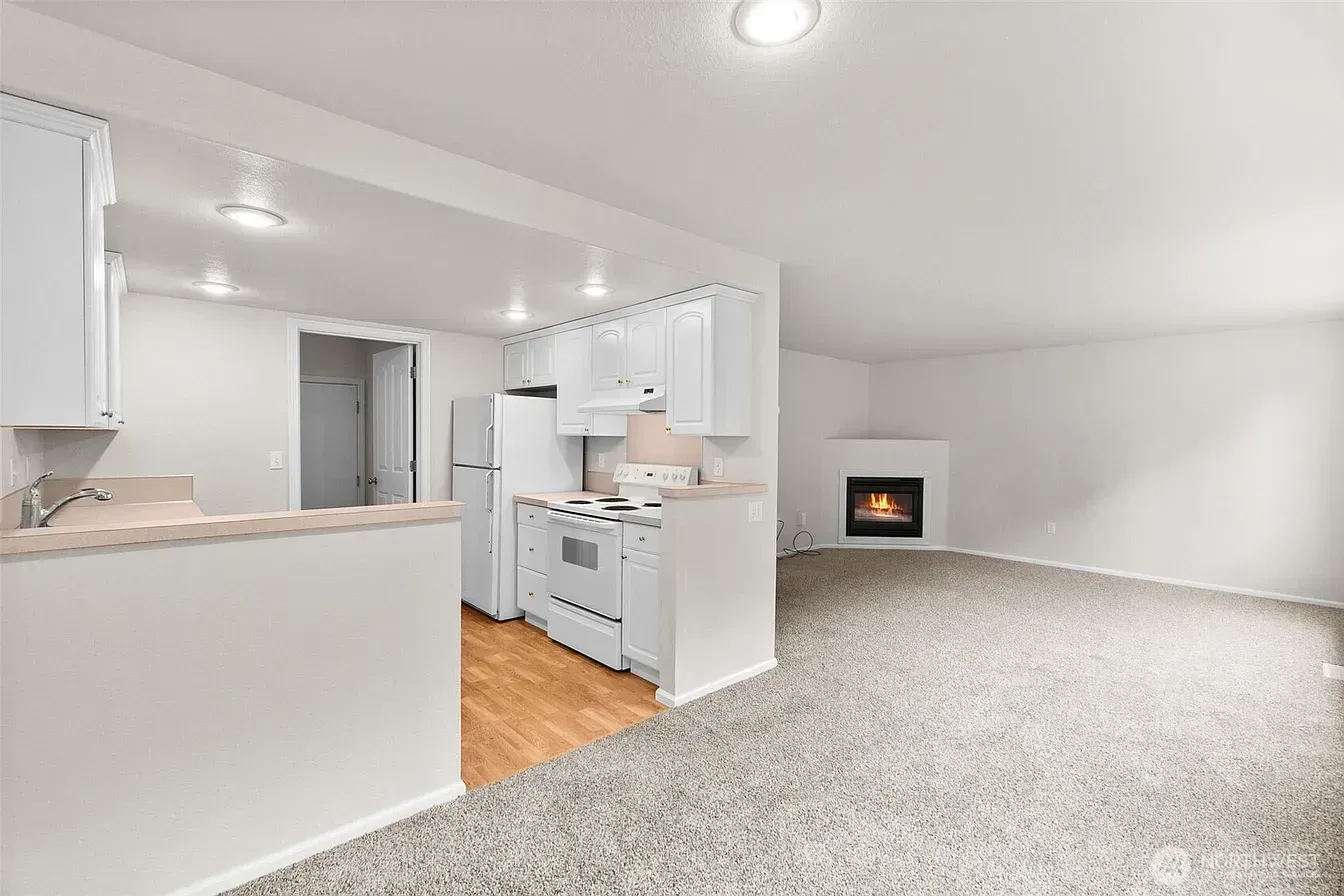 Bright open living room with beige carpet, white kitchen, and small fireplace on far wall