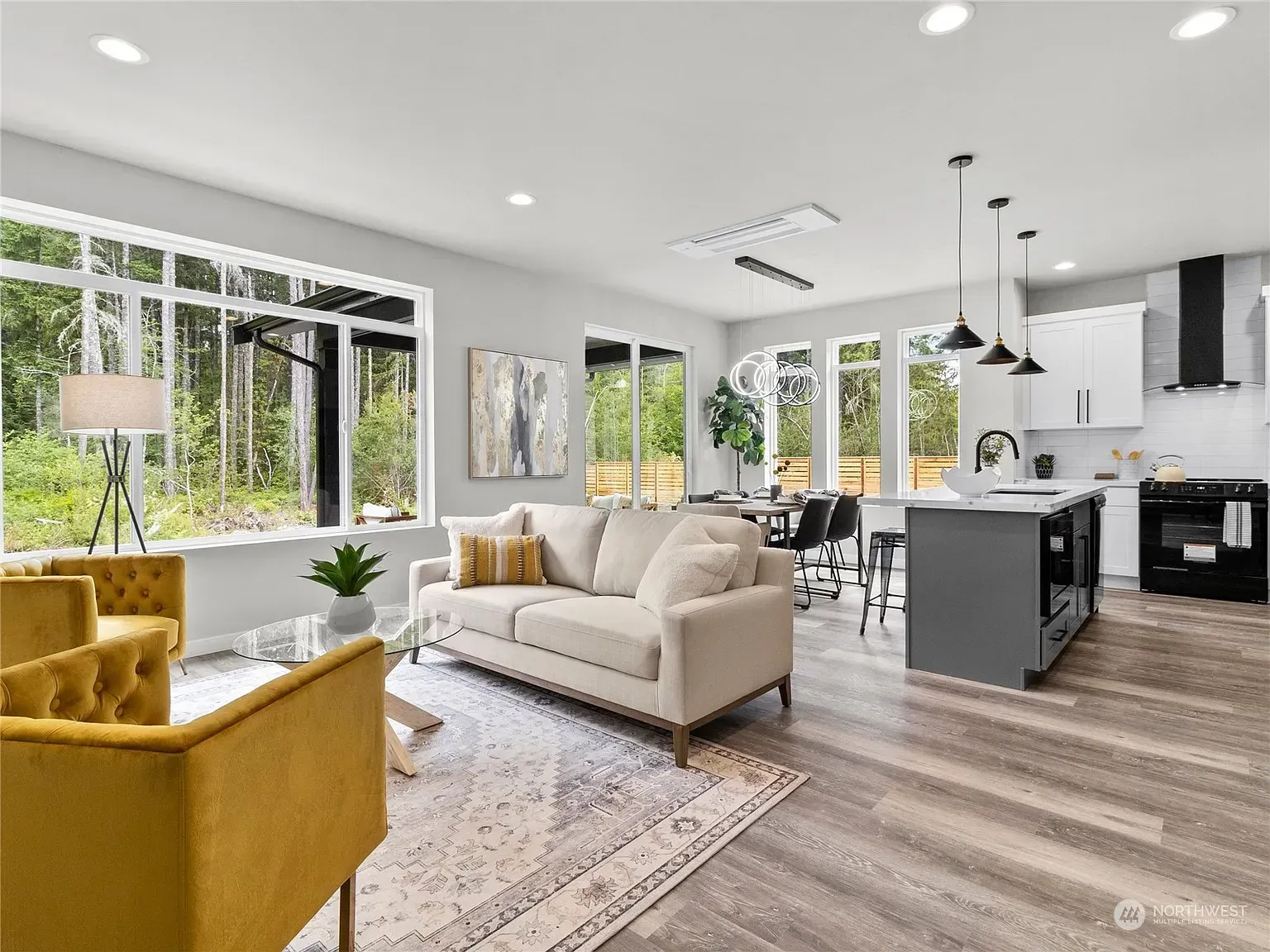 Open-concept living space with beige sofa, yellow chair, kitchen, large windows, and hardwood floors.