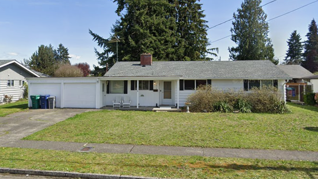 White single-story house with attached garage, green lawn, trees, and blue recycling bins.