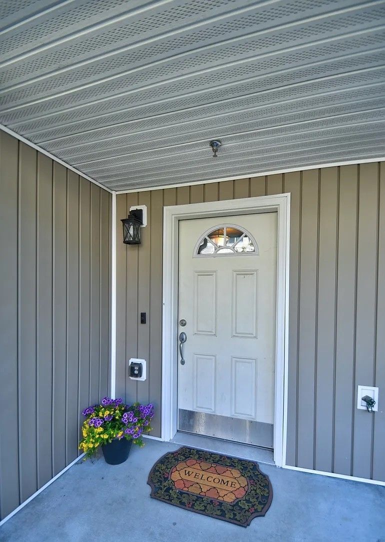 Tan siding and ceiling with a white door, mat, and potted flowers under a covered porch.