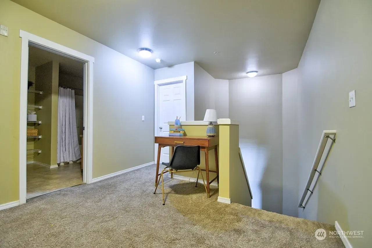 Loft area with desk, chair, and open doorway to a bathroom. Brown carpet and beige walls.