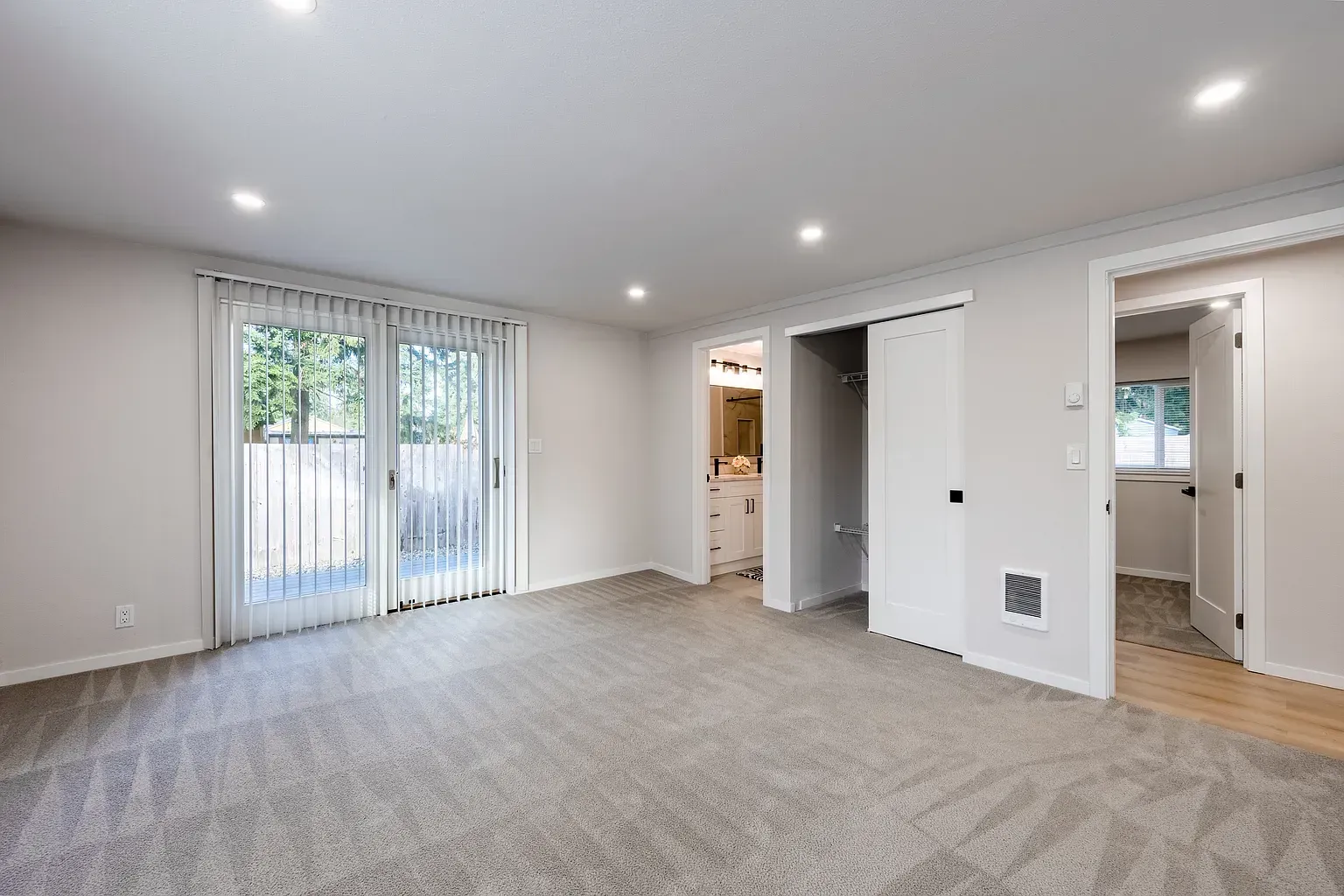 Empty carpeted living room with sliding glass doors, recessed lights, and open doorway to hallway
