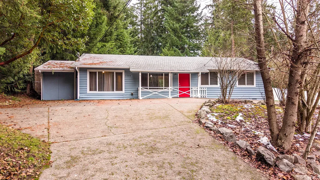 Blue ranch house with red front door, driveway, and surrounding trees.
