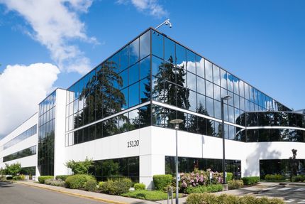 Modern office building with glass facade, trees reflected in windows, sunny day.