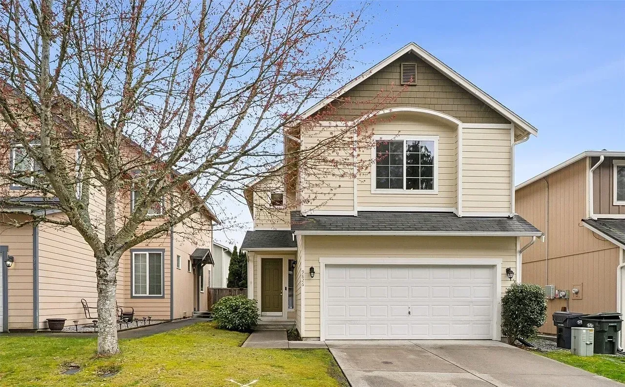 Two-story beige house with a garage and bare tree in front, next to similar houses on a green lawn.